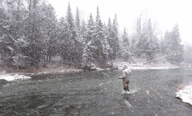 The best waders for winter fishing are boot foot waders - An angler standing in the river and fishing is a snow storm.