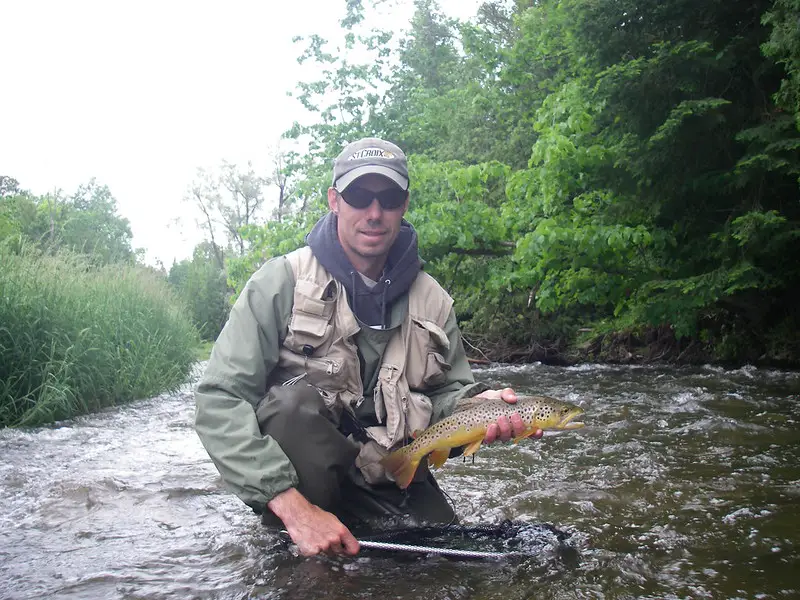 The author with a brown trout in fast flowing river with lots of trees and busk lining the river.